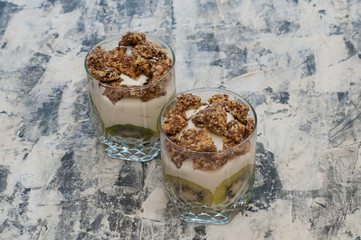 Granola, kiwi, banana and greek yogurt in glass cups on gray concrete