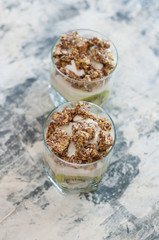 Granola, kiwi, banana and Greek yogurt in two glass cups on gray concrete, close-up view from the top