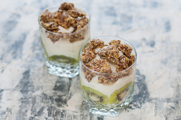 Granola, kiwi, banana and Greek yogurt in two glass cups on gray concrete, copy space. Fitness, figure, body and healthy food