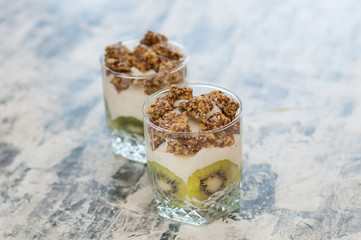 Granola, kiwi, banana and Greek yogurt in two glass cups on gray concrete, copy space. Fitness, figure, body and healthy food