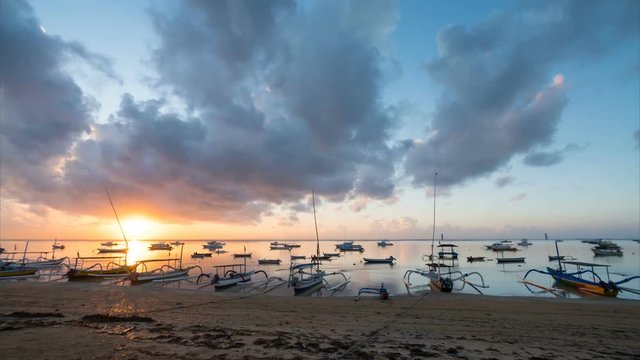 Timelapse of landscape: sun rises above Sanur beach. Amazing cloudscape and traditional jukungs, small wooden Indonesian double outrigger canoes