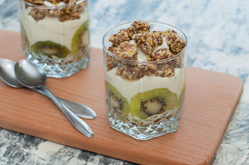 Granola, kiwi, banana and Greek yogurt in two glass cups on wooden board gray concrete, with spoons, copy space. Fitness, figure, body and healthy food