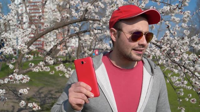 Funny man in red baseball cap in sunglasses with red smartphone listens to music and dances in city park near apricot tree