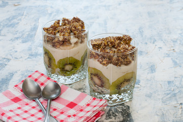Dessert granola, Greek yogurt, kiwi and banana in two glass cups with spoons and pink checkered towel on gray concrete. Fitness, figure, body and healthy food