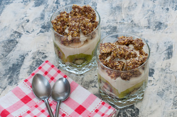 Dessert granola, Greek yogurt, kiwi and banana in two glass cups with spoons and pink checkered towel on gray concrete. Fitness, figure, body and healthy food