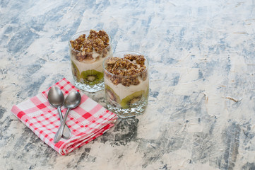 Obraz na płótnie Canvas Dessert granola, Greek yogurt, kiwi and banana in two glass cups with spoons and pink checkered towel on gray concrete. Fitness, figure, body and healthy food