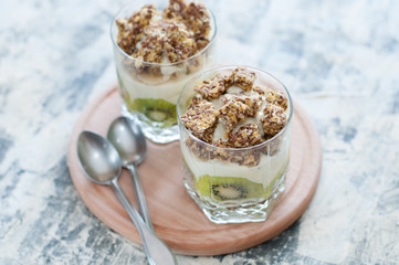 Dessert granola, Greek yogurt, kiwi and banana in two glass cups with spoons on wooden round board, gray concrete, close-up, view from the top. Fitness, figure, body and healthy food