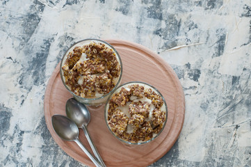 Dessert granola, Greek yogurt, kiwi and banana in two glass cups with spoons on a wooden round board, gray concrete, flat lay. Fitness, figure, body and healthy food