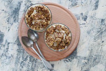 Dessert granola, Greek yogurt, kiwi and banana in two glass cups with spoons on a wooden round board, gray concrete, flat lay. Fitness, figure, body and healthy food