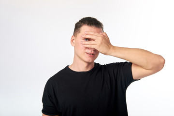 Young attractive curious man hiding his eyes by hand and smiling. Studio shot on white background. I am waiting surprise.
