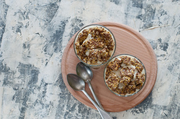 Dessert granola, Greek yogurt, kiwi and banana in two glass cups with spoons on a wooden round...