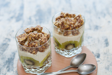 Dessert granola, Greek yogurt, kiwi and banana in two glass cups with spoons on a wooden round board, gray concrete, close-up. Fitness, figure, body and healthy food