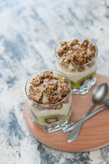 Dessert granola, Greek yogurt, kiwi and banana in two glass cups with spoons on a wooden round board, gray concrete, close-up. Fitness, figure, body and healthy food