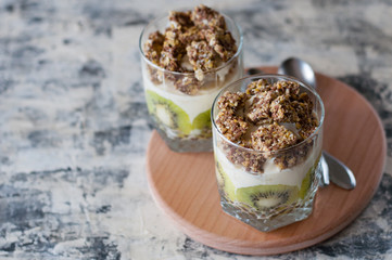Dessert granola, Greek yogurt, kiwi and banana in two glass cups with spoons on a wooden round board, gray concrete, copy space. Fitness, figure body and healthy food