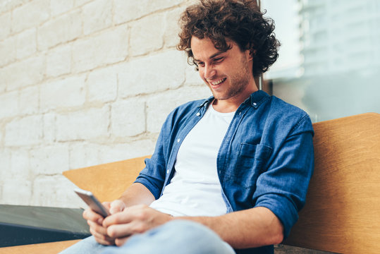 Young man with curly hair stitting outdoors typing on mobile phone. Happy male using smart phone application for searching location in downtown in the city. Lifestyle, technology and people concept