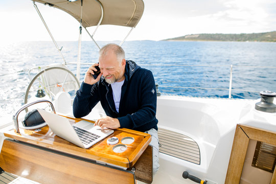 Digitaler Nomade arbeitet am Laptop lachend und telefonierend auf einem Segelboot