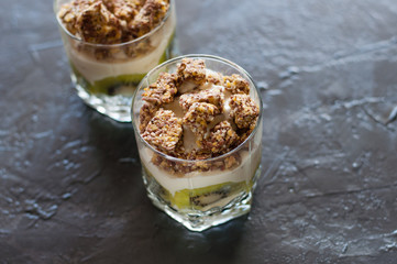 Greek yogurt, granola, banana and kiwi. Two desserts in glass cups on dark background. Concept body and healthy food