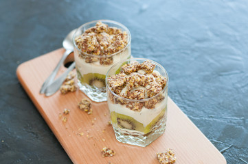 Greek yogurt, granola, banana and kiwi. Desserts in glass cups with spoons on wooden platform, dark background, view from the top. Concept body and healthy food