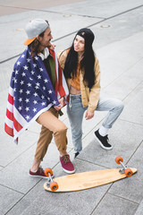 high angle view of man with american flag on shoulders looking at brunette woman, standing near skateboard
