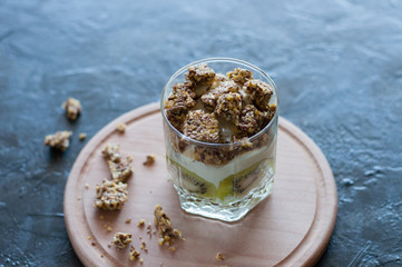 Granola, banana, kiwi and greek yogurt. Dessert in glass cup on a wooden round board, dark background. Concept body and healthy food