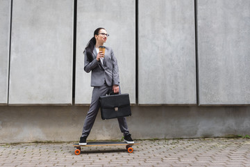 Cheerful businesswoman in formal wear riding on skateboard, holding paper cup and briefcase in hands