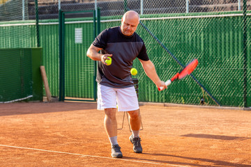 A middle-aged bald man plays tennis on the outdoor court. Sunny day.