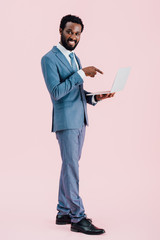 smiling african american businessman pointing at laptop isolated on pink