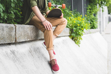 cropped view of man in casual wear holding skateboard in hand, sitting on border