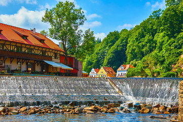Cesky Krumlov (Czech Krumlov), Czech Republic. Antique town on river Vltava. Picturesque landscape. Cascade dam with stones. Colourful houses on bank among green trees. Sunny summer day with blue sky.