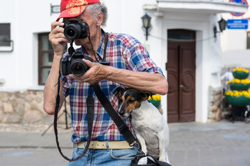 The old man on the move takes pictures at once on two cameras. A faithful dog sits on his master's bag and looks at. funny photo