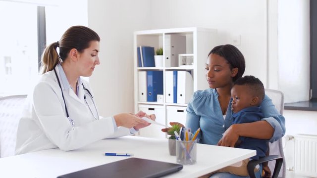 medicine, healthcare and pediatry concept - caucasian doctor giving prescription to african american mother with baby son at clinic