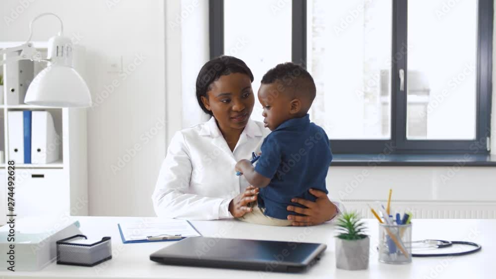 Wall mural medicine, healtcare and pediatry concept - happy african american female doctor or pediatrician holding baby boy patient on medical exam at clinic