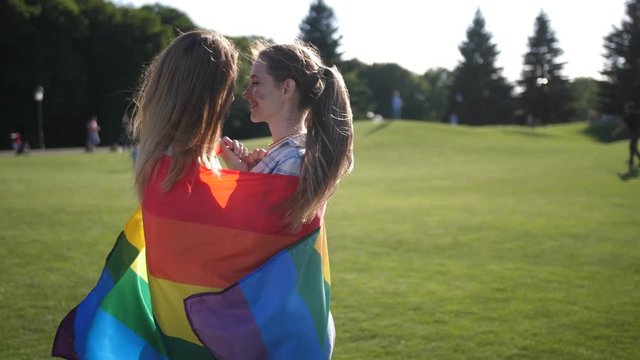 Close-up of attractive same-sex couple with rainbow flag painted on cheeks rubbing noses standing close in the middle of park lawn. Summer wind fluttering color lgbt flag and waving light women's hair