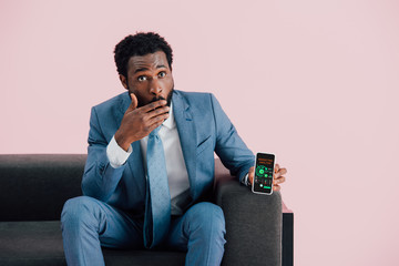 shocked african american businessman in suit sitting on armchair and showing smartphone with...