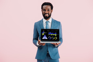 smiling african american businessman showing laptop with infographic isolated on blue