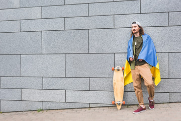 happy man in ukrainian flag on shoulders looking away, standing near wall with skateboard