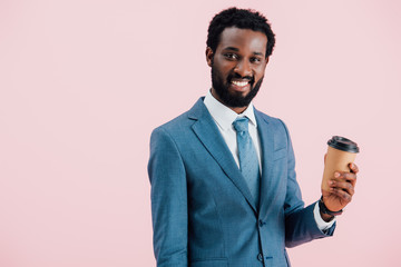 happy african american businessman holding coffee to go, isolated on pink