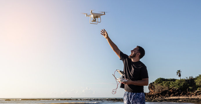 Using a drone uses remote control on the beach. Full length shot of young man operating the drone by remote control at the sea shore. Brazil.