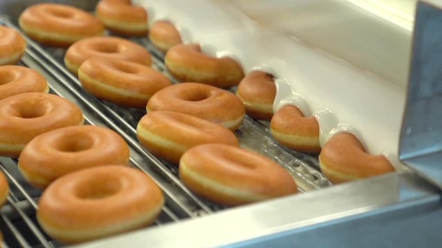 This video shows a busy donut factory adding icing to hot, fresh glazed donuts on a production conveyor belt.