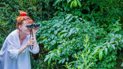 A curious woman is standing in a garden between plants and secretly observes. She is holding...