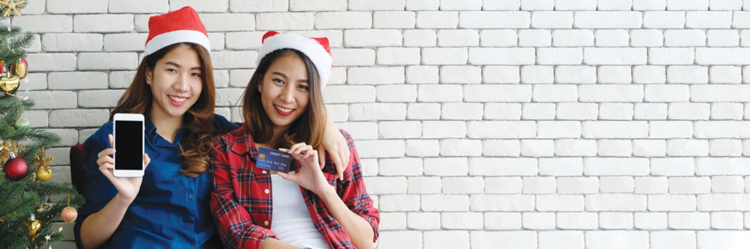 Two young cute asia women holding smartphone with blank screen and credit card for shopping online background, banner, mock up, with happiness, Christmas holiday shopping concept