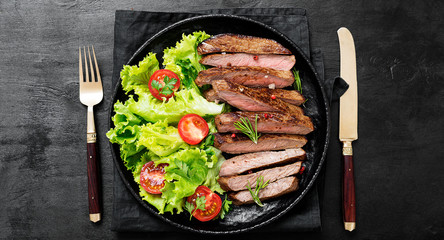 Sliced grilled beef steak and green salad leaves on black plate.Top wiev.