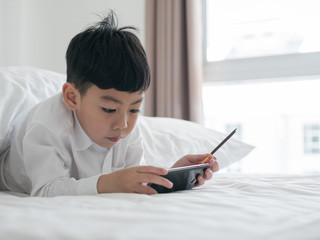 Cute little asian kid focused on smartphone while lying on the bed. Boy playing games on smartphone. Social and technology concept