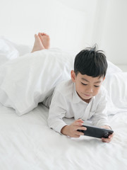 Cute little asian kid focused on smartphone while lying on the bed. Boy playing games on smartphone. Social and technology concept