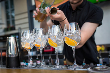 four glasses standing on the bar. Glasses are with ice and orange. Barman pours alcohol in glasses from a measuring glass