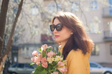 Side view of beautiful cheerful girl with cute sensual lips wearing sunglasses. Young woman in yellow jacket holding a big bouquet of colorful flowers outdoors near the buildings on the background