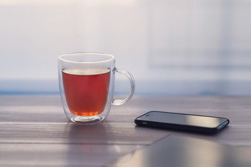 Transparent cup of tea, phone, tablet on a white wooden table.