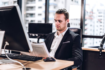 Naklejka na ściany i meble Confident young handsome businessman reading newspaper and working with computer in modern office