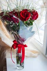 Lovely big bouquet with flowers of roses of vinous red color are standing in the glass vase on the table. Green leaves and thorns. Wide red bow. Calm light background