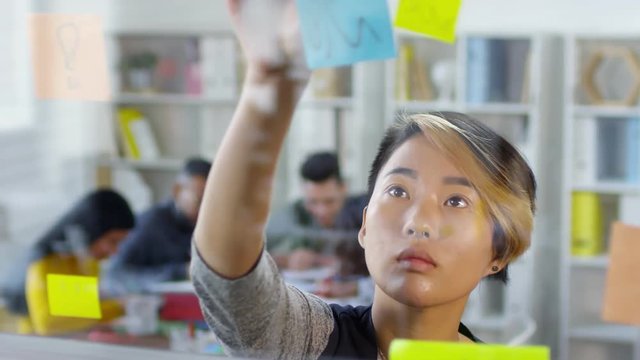 Close up shot of young Asian female employee with creative hairstyle standing in front of glass wall in office and putting up post it reminders